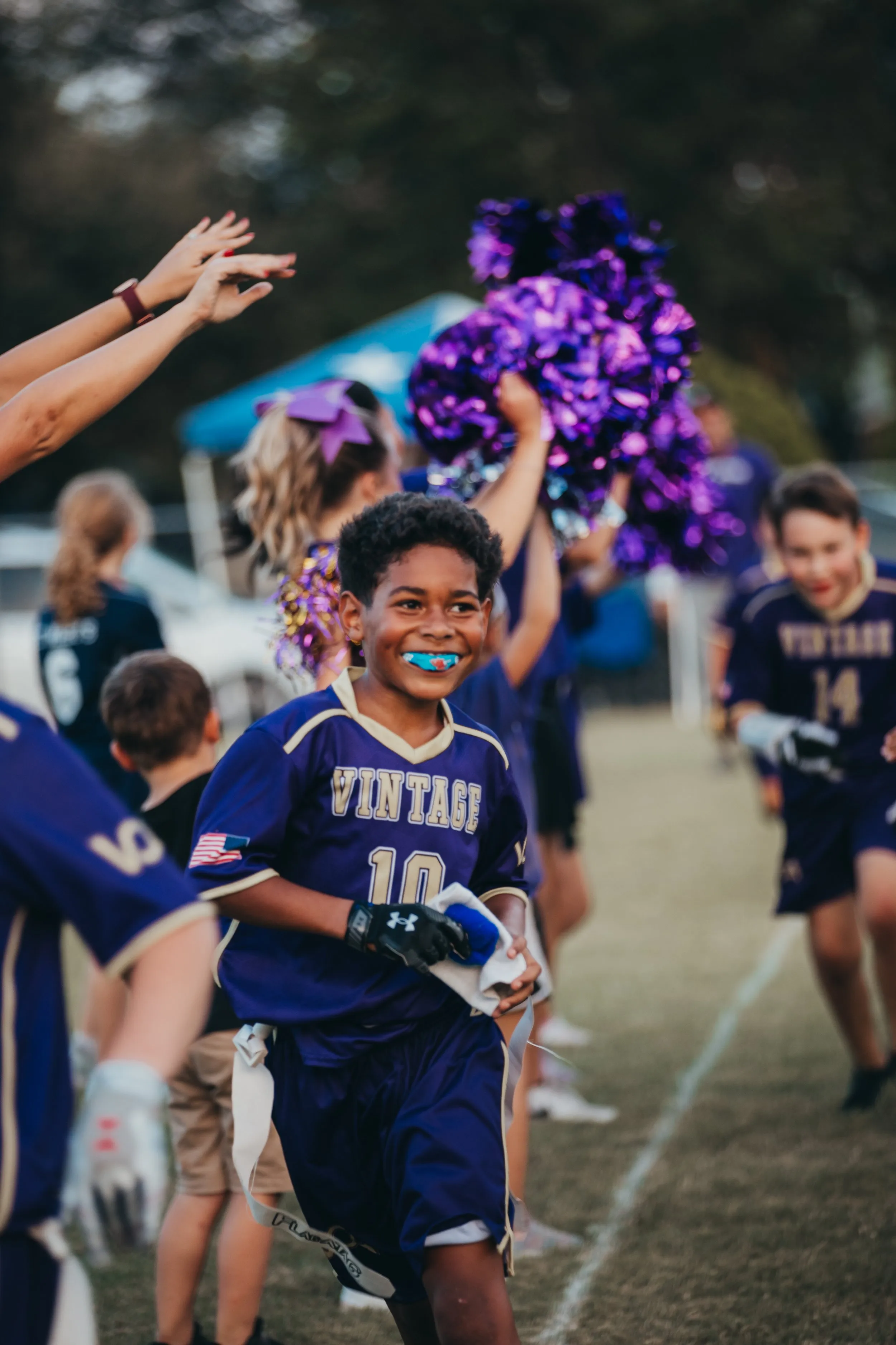 A young Vintage Warriors flag football player smiles running onto the field as teammates and cheerleaders look on.