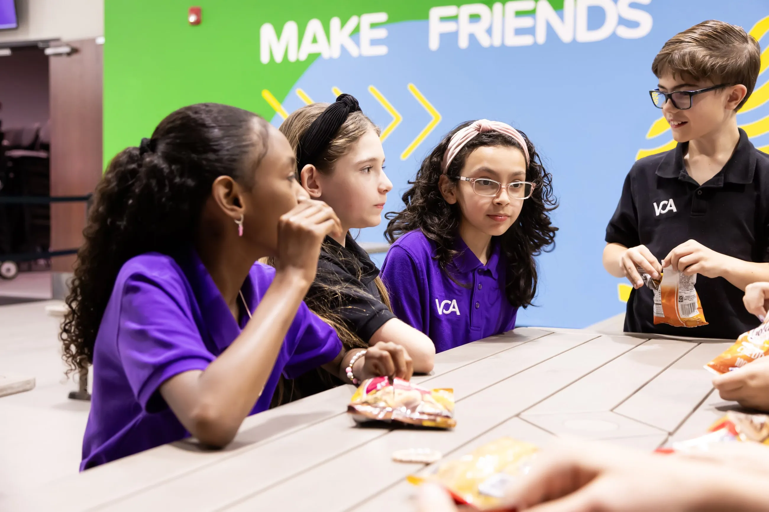 Students share snacks and conversation at a table beneath a colorful “Make Friends” mural.