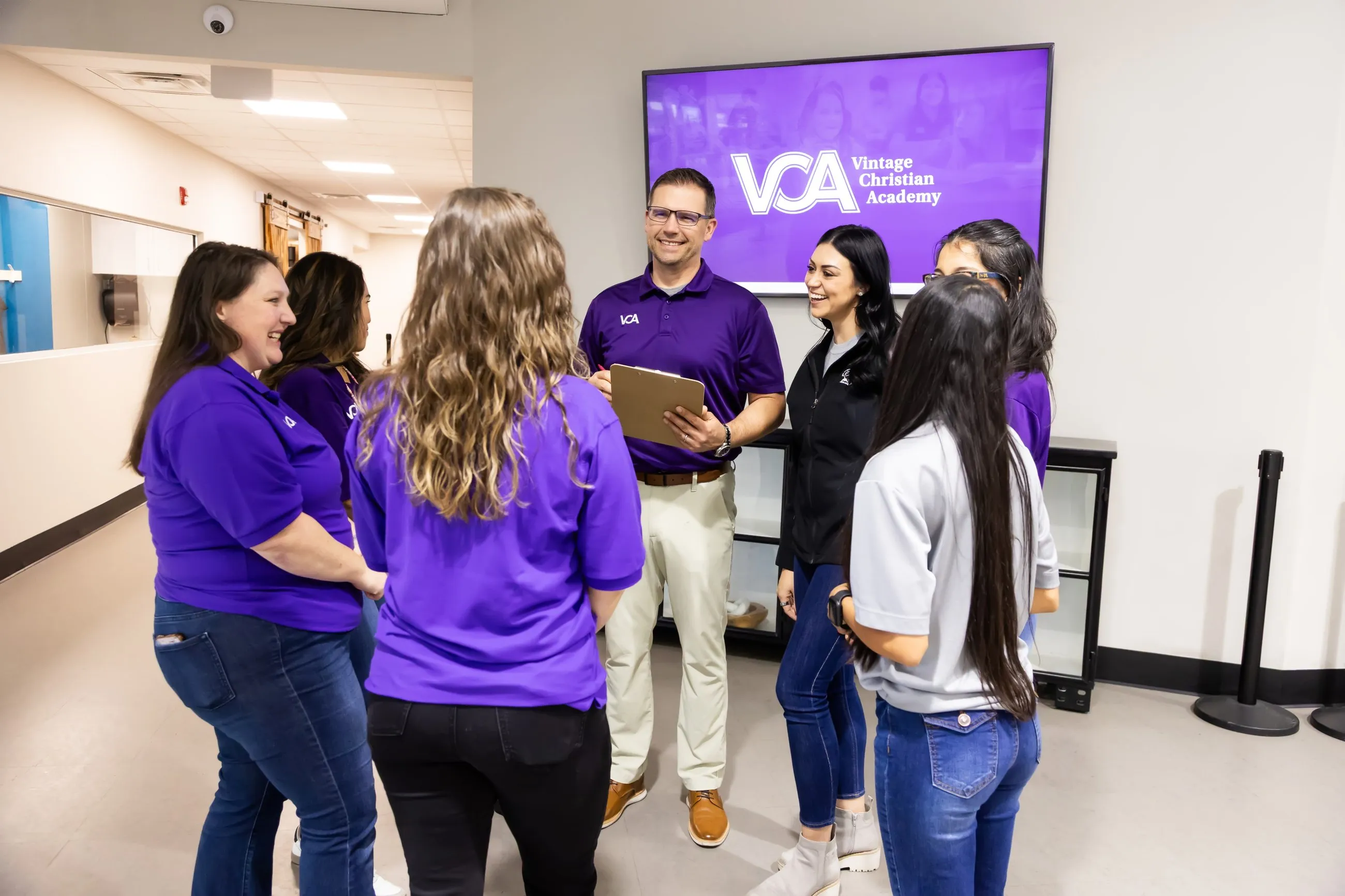 Staff members gather and talk warmly in the school hallway beneath a screen displaying the Vintage Christian Academy logo.