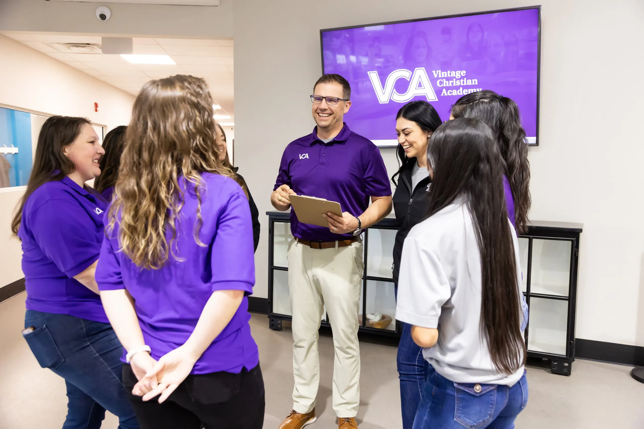 Staff members share a warm conversation in the school hallway beneath a screen displaying the Vintage Christian Academy logo.