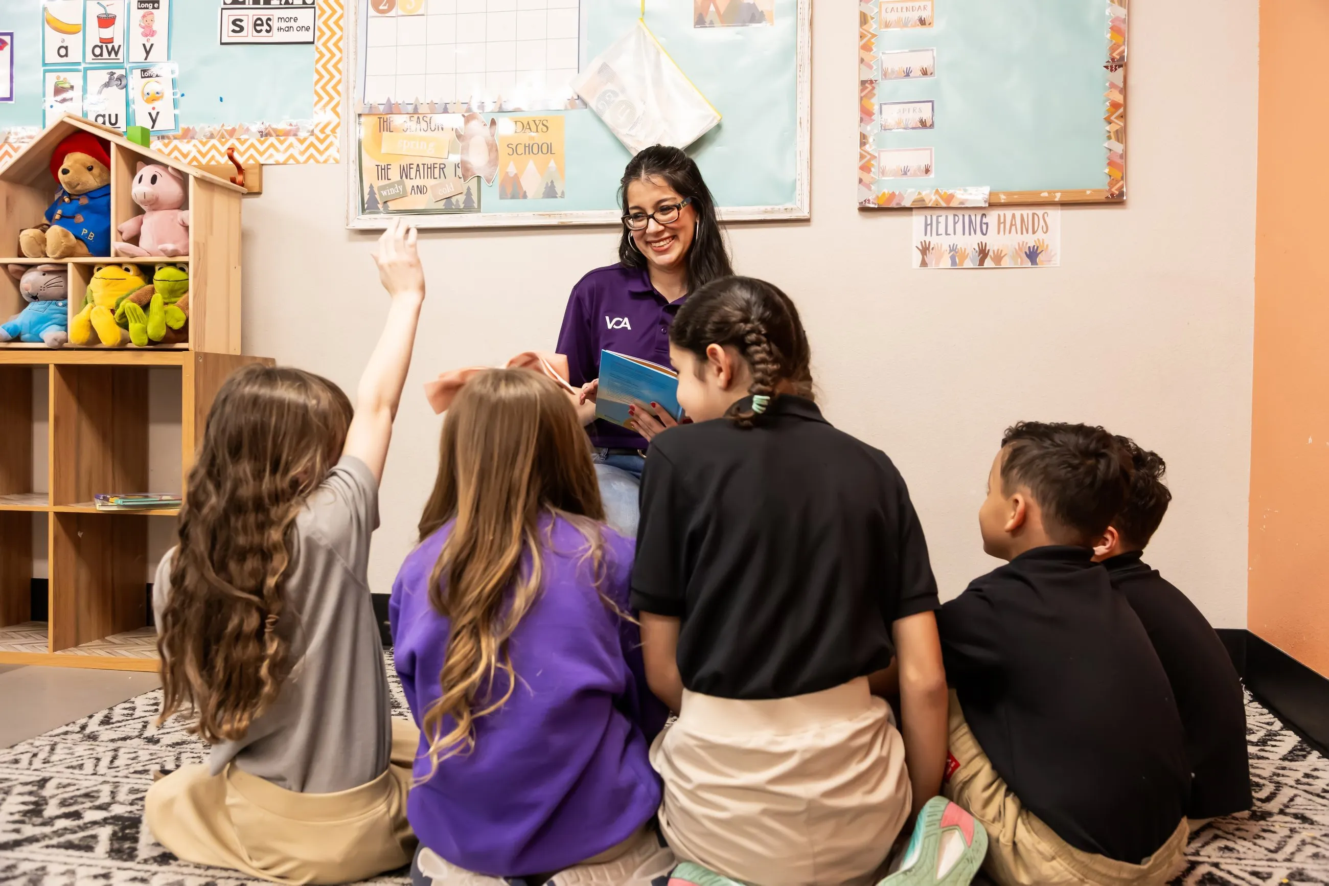 A teacher reads aloud to young students seated on the classroom rug, one raising her hand.