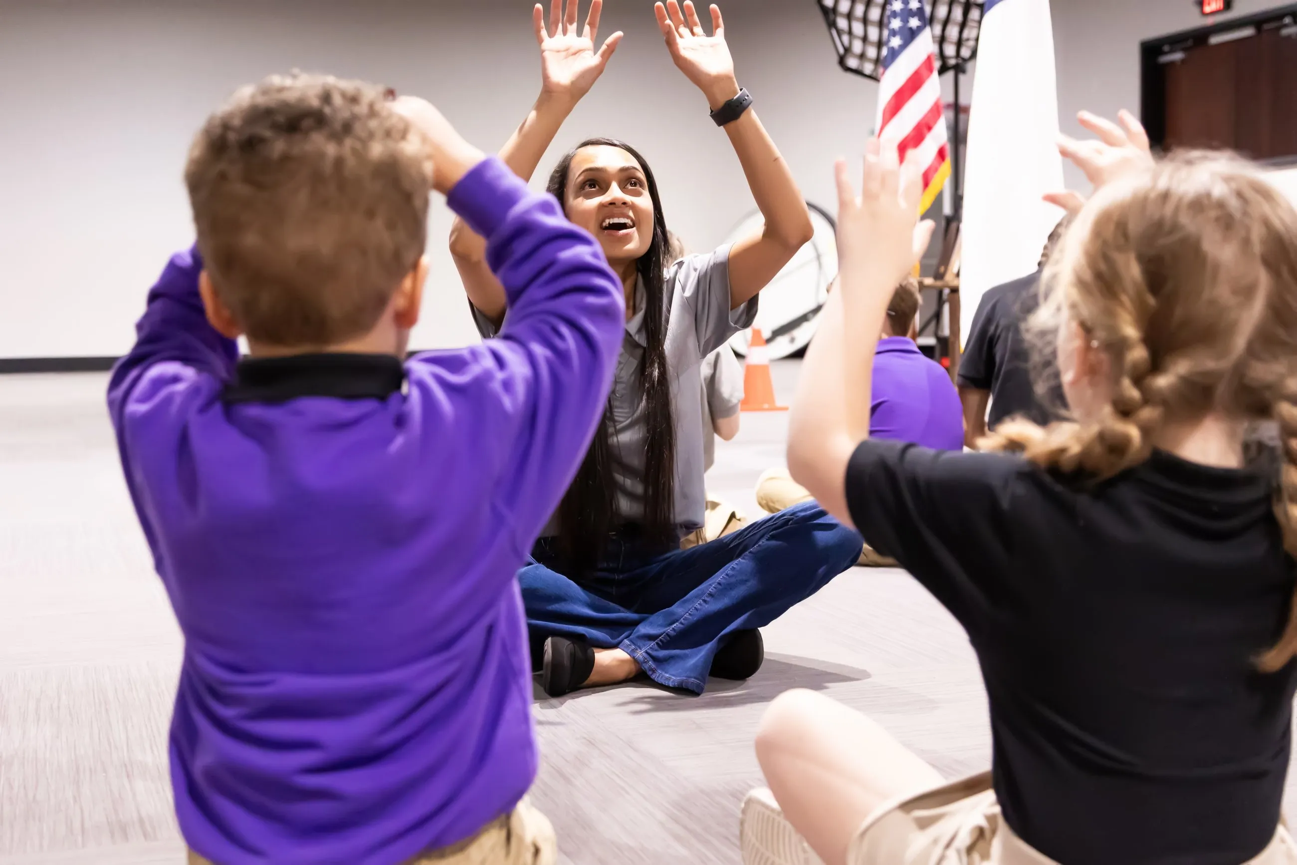 A teacher sits on the floor with students, hands raised together during a classroom activity.